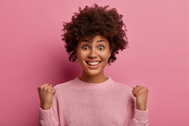 Excited cheerful woman smiles broadly, awaits important results, rejoices positive news, achieves goal, looks happily , wears casual sweater, poses over rosy wall, going to grab opportunity