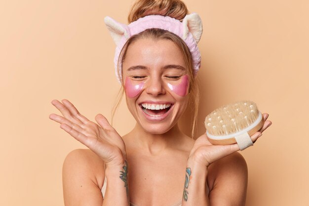 Excited cheerful woman smiles broadly keeps eyes closed applies pink hydrogel patches holds bath brush enjoys spa procedures at home stands topless against beige background. Wellness and beauty