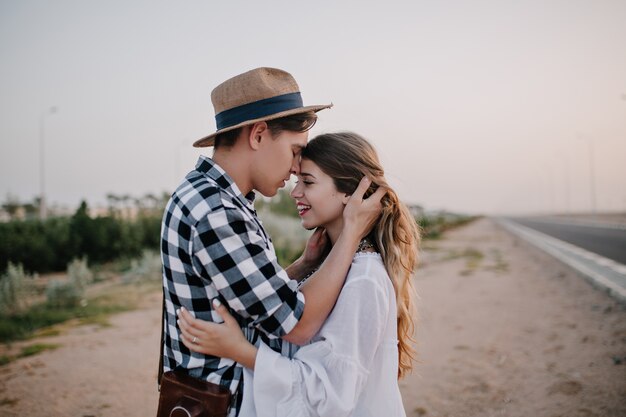 Excited cheerful woman in white tunic soft embracing boyfriend in stylish shirt, while he touching her long hair. Young couple of travelers hugging standing near the road and enjoys evening date