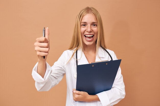 Excited cheerful young woman physician with braces and blonde hair posing holding clipboard and pen making thumbs up gesture, opening mouth broadly