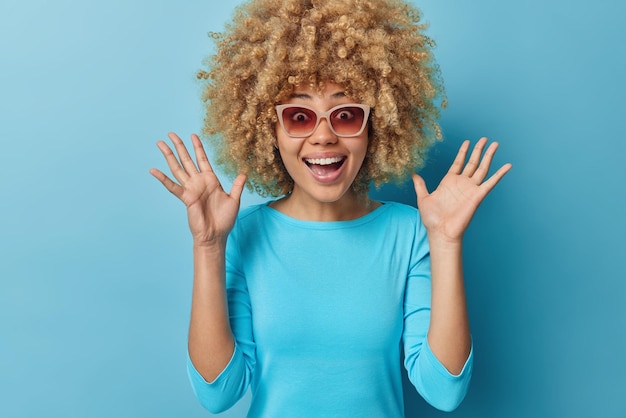 Foto gratuita eccitata giovane donna allegra con i capelli ricci tiene i palmi delle mani alzati esclama felicemente indossa occhiali da sole maglione casual isolato su sfondo blu reagisce su un'offerta straordinaria concetto di emozioni umane