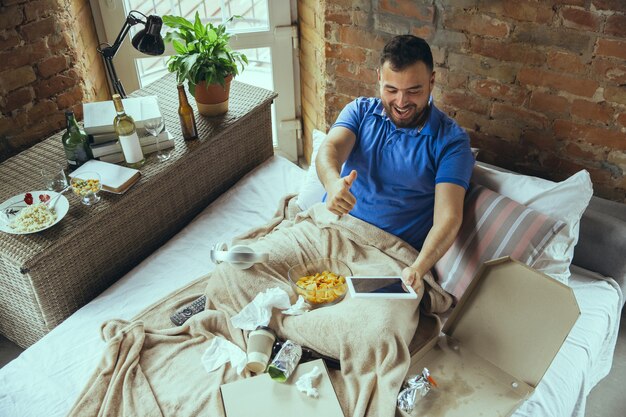 Excited cheering of sport team, thumb up. Lazy man living in his bed surrounded with messy. No need to go out to be happy. Using gadgets, watching movie and series, emotional. Fast food.