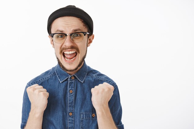 Excited cheering unshaven male model feeling happy and triumphing raising clenched fists and yelling supportive smiling broadly