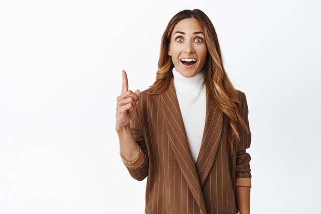 Excited creative businesswoman got an idea found solution pointing finger up and smiling amazed eureka gesture standing in business suit over white background