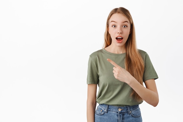 Excited and curious girl pointing left with opened mouth, showing awesome new promo deal, store banner on copyspace, standing over white wall