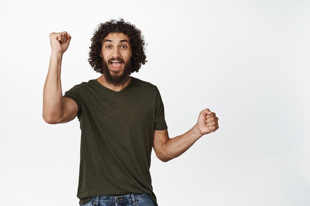 Excited curly guy chanting celebrating victory raising hands up and triumphing standing in tshirt over white background Copy space