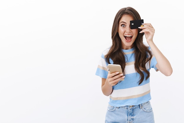 Excited cute good-looking woman in t-shirt, jeans, hold smartphone and cover one eye credit card, smiling amused