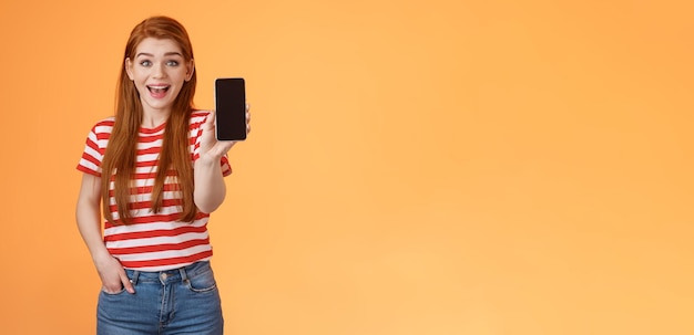 Foto gratuita eccitata donna rossa uscente carina impressionata mostrando l'app tenere lo smartphone introduce la funzione gadget