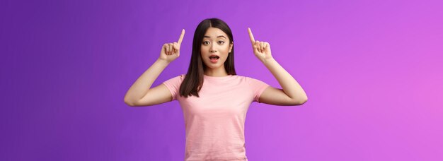 Excited cute surprised asian girl in pink tshirt telling about awesome news pointing raised fingers