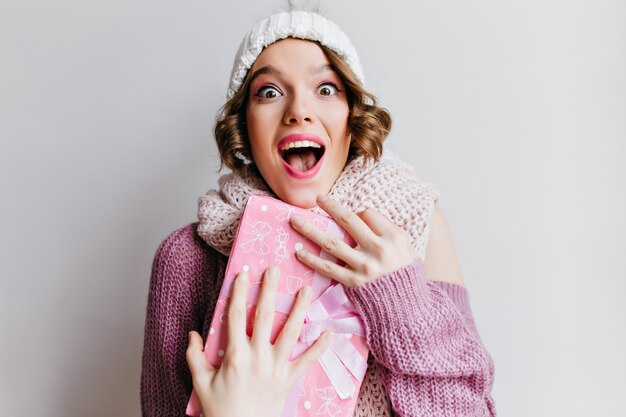 Excited dark-eyed woman in warm hat posing emotionally on white wall. Indoor photo of surprised beautiful girl in winter accessories.