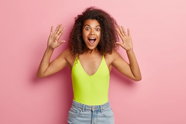 Excited dark skinned Afro woman raises palms, wears bright top and denim shorts, keeps mouth widely opened