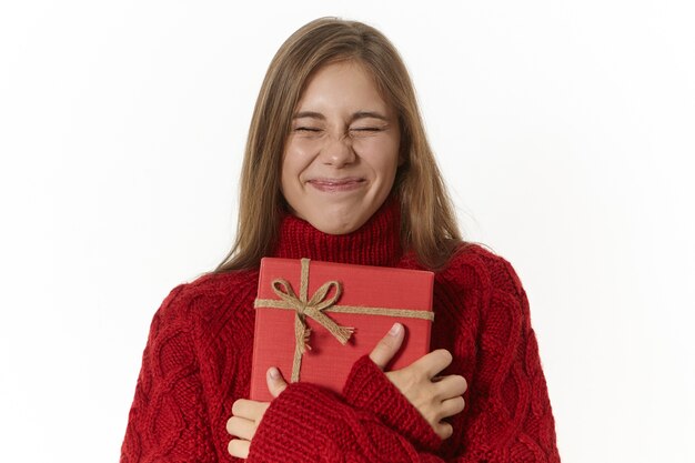 excited ecstatic girl in cozy knitted sweater keeping eyes closed and embracing fancy red box, feeling impatient to open it