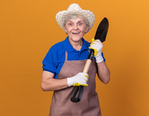 Excited elderly female gardener wearing gardening hat and gloves holding spade isolated on orange wall with copy space