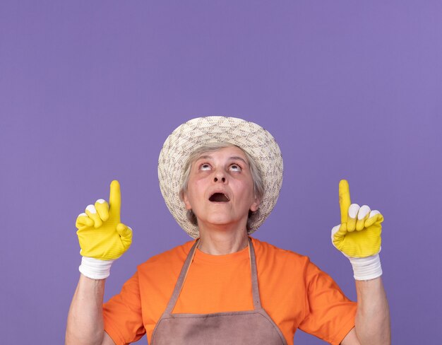 Excited elderly female gardener wearing gardening hat and gloves looking and pointing up