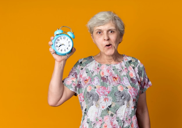 Возбужденная пожилая женщина держит будильник на оранжевой стене