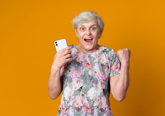 Excited elderly woman raises fist holding phone isolated on orange wall