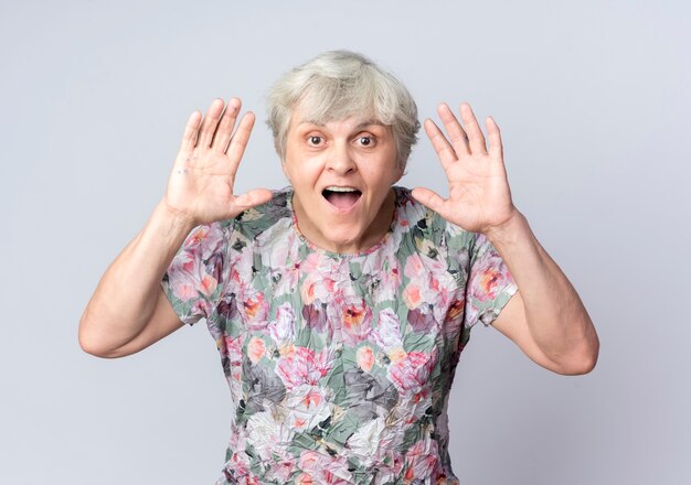 Excited elderly woman stands with raised hands isolated on white wall