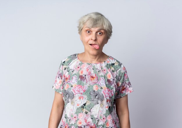 Excited elderly woman sticks tongue out isolated on white wall