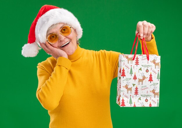 Возбужденная пожилая женщина в солнцезащитных очках в шляпе санта-клауса кладет руку на лицо и держит бумажный подарочный пакет, изолированный на зеленом фоне с копией пространства