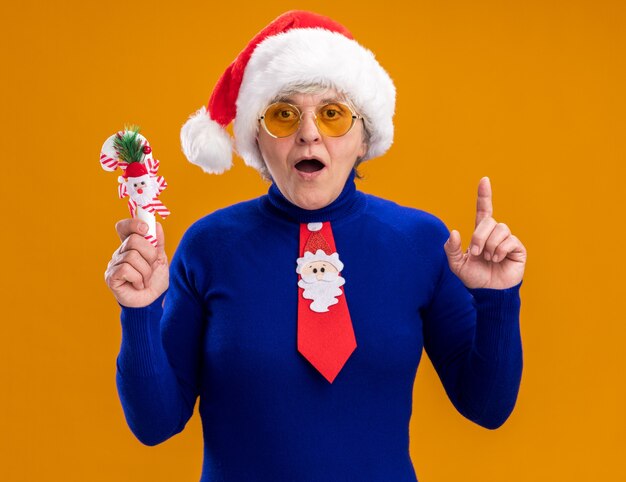 excited elderly woman in sun glasses with santa hat and santa tie holds candy cane and points up isolated on orange background with copy space