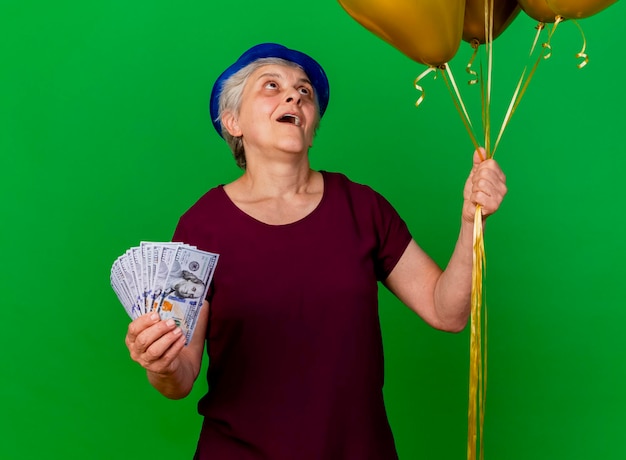 Excited elderly woman wearing party hat holds money and looks at helium balloons on green