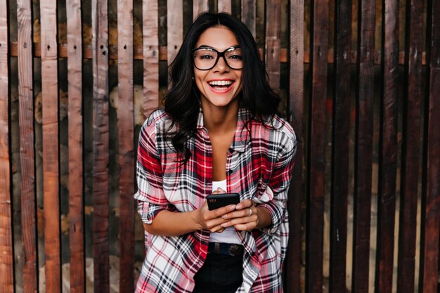 Excited elegant girl in spring outfit laughing on wooden wall. Outdoor shot of amazing latin woman in glasses holding phone with smile.