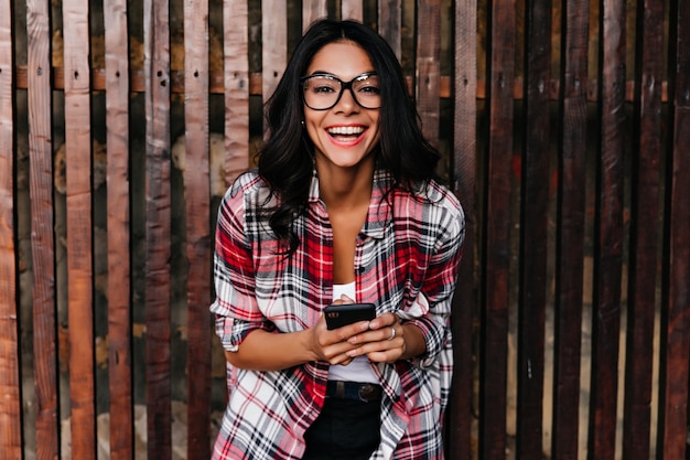 木製の壁で笑っている春の衣装で興奮したエレガントな女の子。笑顔で携帯電話を保持しているメガネで素晴らしいラテン女性の屋外ショット。