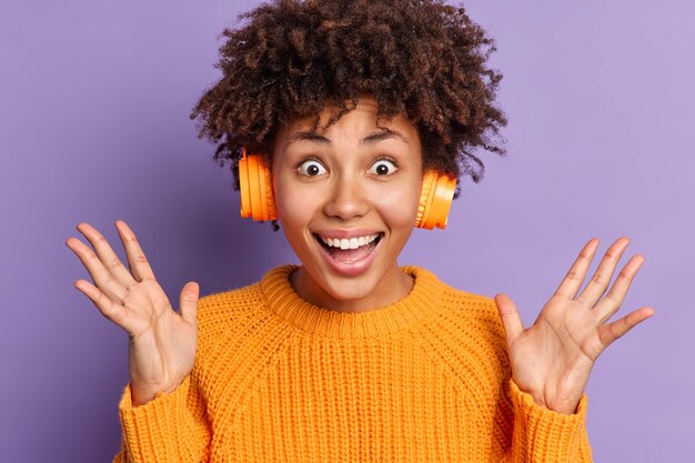 Excited emotional Afro American woman raises hands reacts happily on amazing news listens favorite music via headphones wears casual orange jumper 