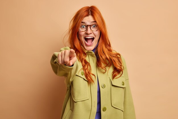 Excited emotional redhead girl with surprised expression exclaims loudly and points directly picks or chooses you dressed in fashionable clothes.