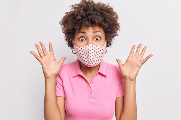 Excited emotional woman stares bugged eyes raises palms wears protective disposable mask to prevent coronavirus spread stays on self isolation during quarantine isolated on white wall