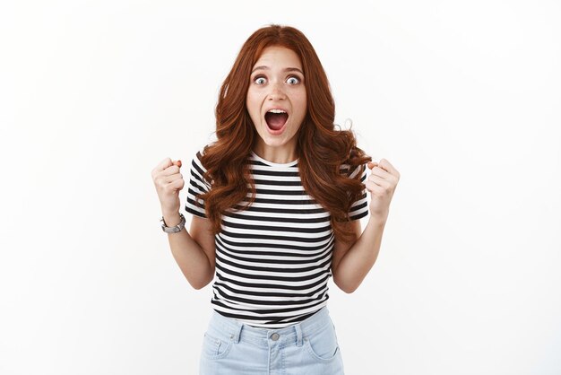 Excited emotive and thrilled gorgeous redhead girl in striped tshirt stare camera surprised and amazed gasping drop jaw fantastic news fist pump triumphing achieve success win prize