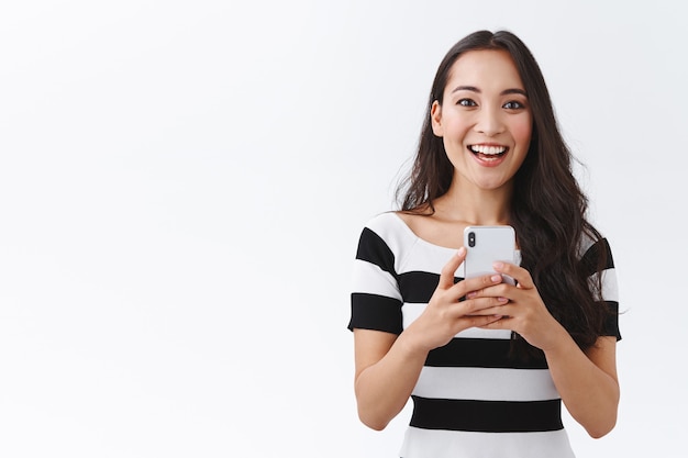 Excited, enthusiastic good-looking asian woman in casual outfit holding smartphone, thrilled smiling and looking camera, record video or photographing via mobile phone, white background