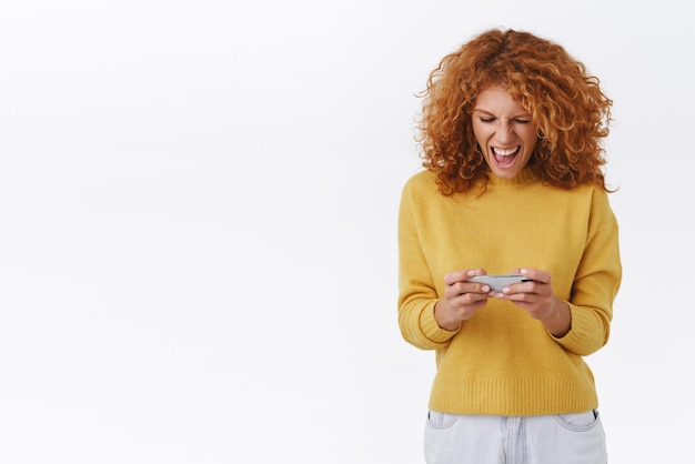 Взволнованная восторженная симпатичная рыжеволосая кудрявая девушка, гримасничая, играя в интенсивную онлайн-игру с помощью мобильного устройства, держа смартфон горизонтально, крича и глядя на экран телефона на белом фоне