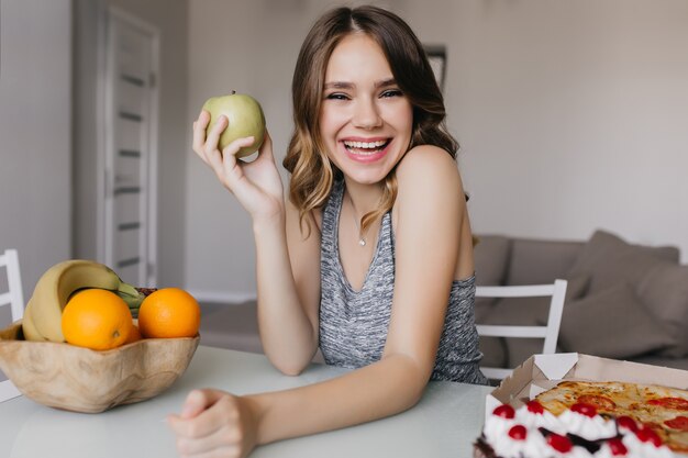 Excited european girl enjoying tasty fruits during diet. Pretty female model holding apple and smiling