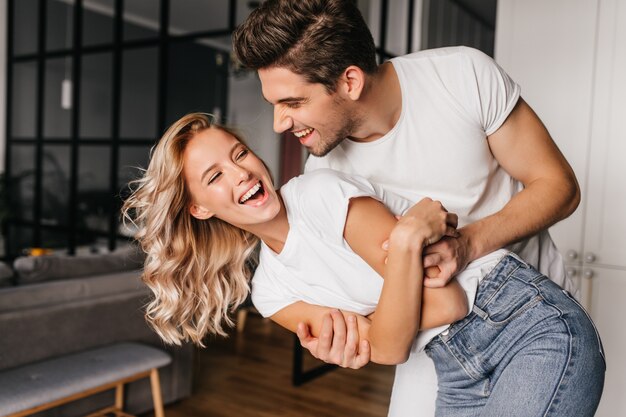 Excited fair-haired girl fooling around with boyfriend at home. Good-humoured brunette man dancing with wife.