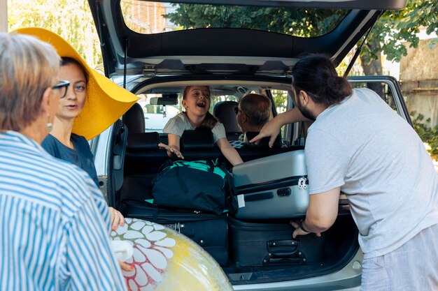 Возбужденная семья едет в отпуск на машине, кладя тележку и сумки в багажник автомобиля. Родители, бабушка и дедушка уезжают в отпуск с маленькой девочкой, пакуя чемодан и багаж.