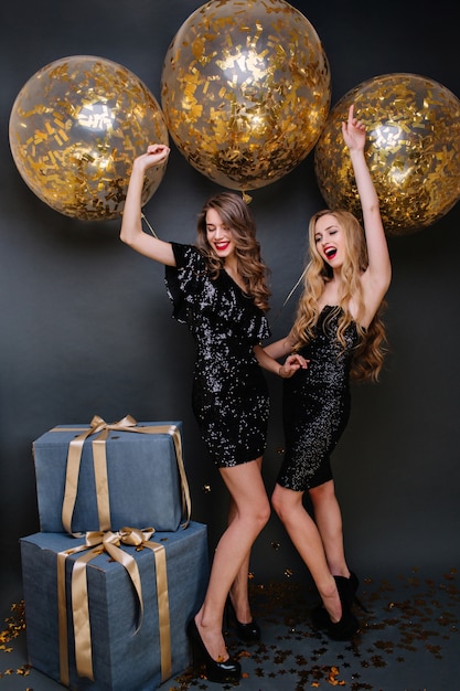 金色のティンセルが付いた大きな風船で新年のパーティーを祝う豪華な黒のドレスを着た興奮したファッショナブルな若い女性。楽しんで、プレゼントして、積極性を表現してください。