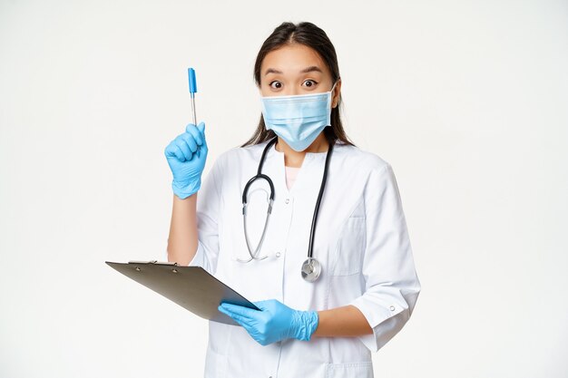 흥분한 여의사, 클립보드를 들고 펜을 들고 있는 아시아 의사, 흰색 배경 위에 의료용 얼굴 마스크를 쓴 채 해결책이나 아이디어를 찾았습니다.