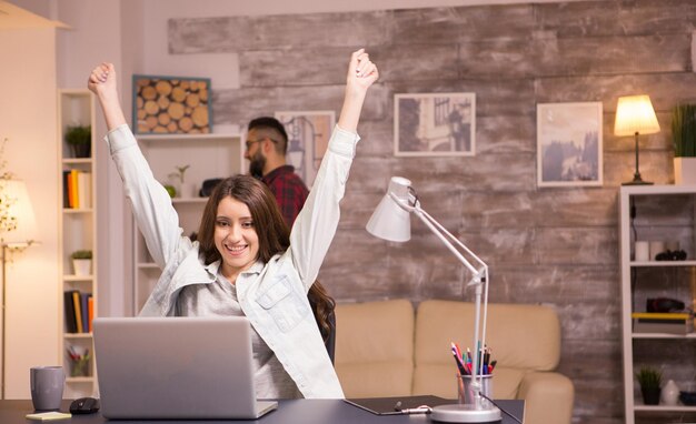 Взволнованная женщина-фрилансер с поднятыми руками после чтения хороших новостей на ноутбуке. Парень разговаривает по телефону в фоновом режиме.