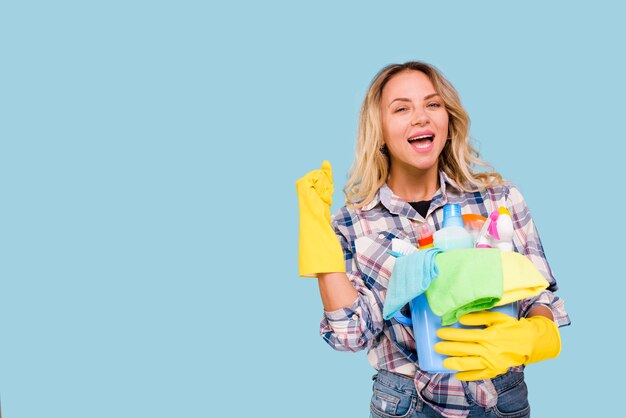 Excited female housekeeper holding bucket with cleaning products while clenching her fist over colored background