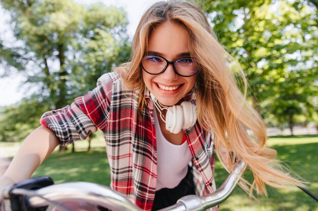 Excited female model in glasses and headphones riding around park. Emotional blonde girl sitting on bicycle and laughing.