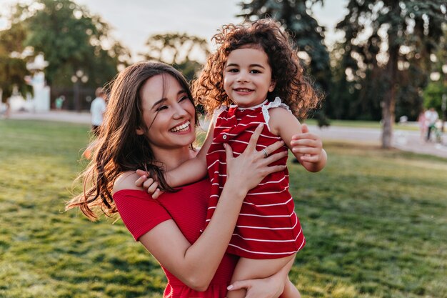 Excited female model holding little girl and smiling on nature  Outdoor photo of spectacular white woman fooling around with daughter during rest in park.