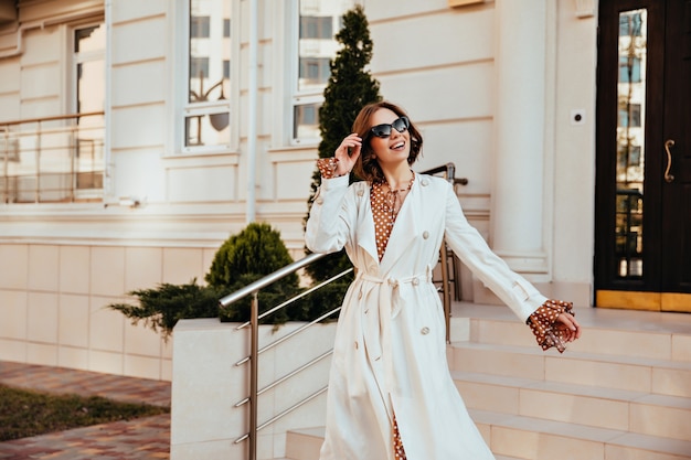 Взволнованная женская модель в длинном белом халате наслаждается добрым днем. Открытый выстрел активной молодой женщины в осеннем наряде.