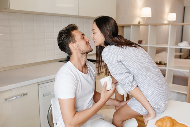 Excited female model sits on table kissing man in white t-shirt, enjoying good morning