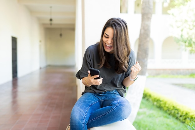 Excited female raising fist and cheering while watching sport on mobile phone in campus