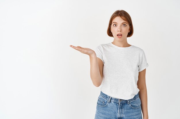 Excited female student holding empty space on palm, holding something in hand and look amazed , standing on white wall