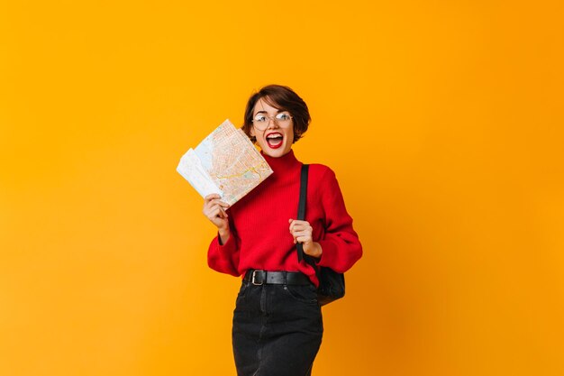 黄色の背景に立っている赤いセーターで興奮した女性旅行者地図と女性のスタジオショット