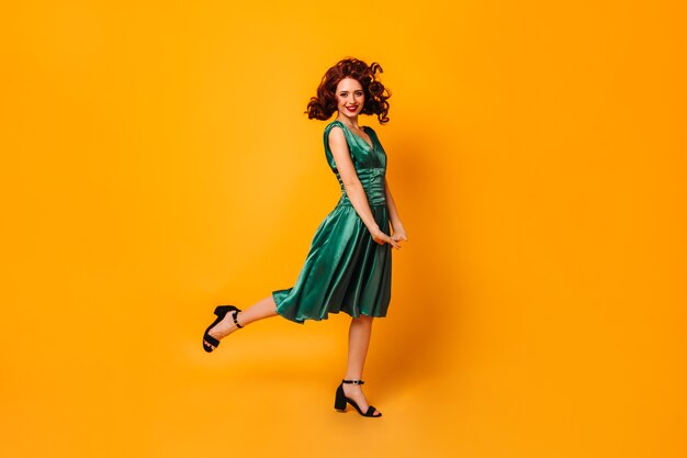 한쪽 다리에 서있는 녹색 드레스에 흥분된 맞는 여자. 노란색 공간에서 춤추는 우아한 아가씨의 전체 길이보기.