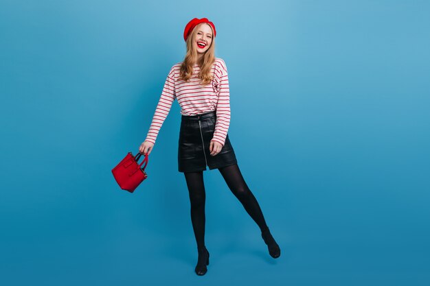 Excited french girl in short skirt dancing on blue wall. Full length view of amazing blonde woman holding red handbag.
