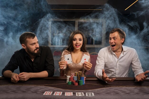 Excited friends are playing poker at casino in smoke, rejoicing in winning and looking at the camera. Youth are making bets waiting for a huge win. Gambling for money. Games of fortune.